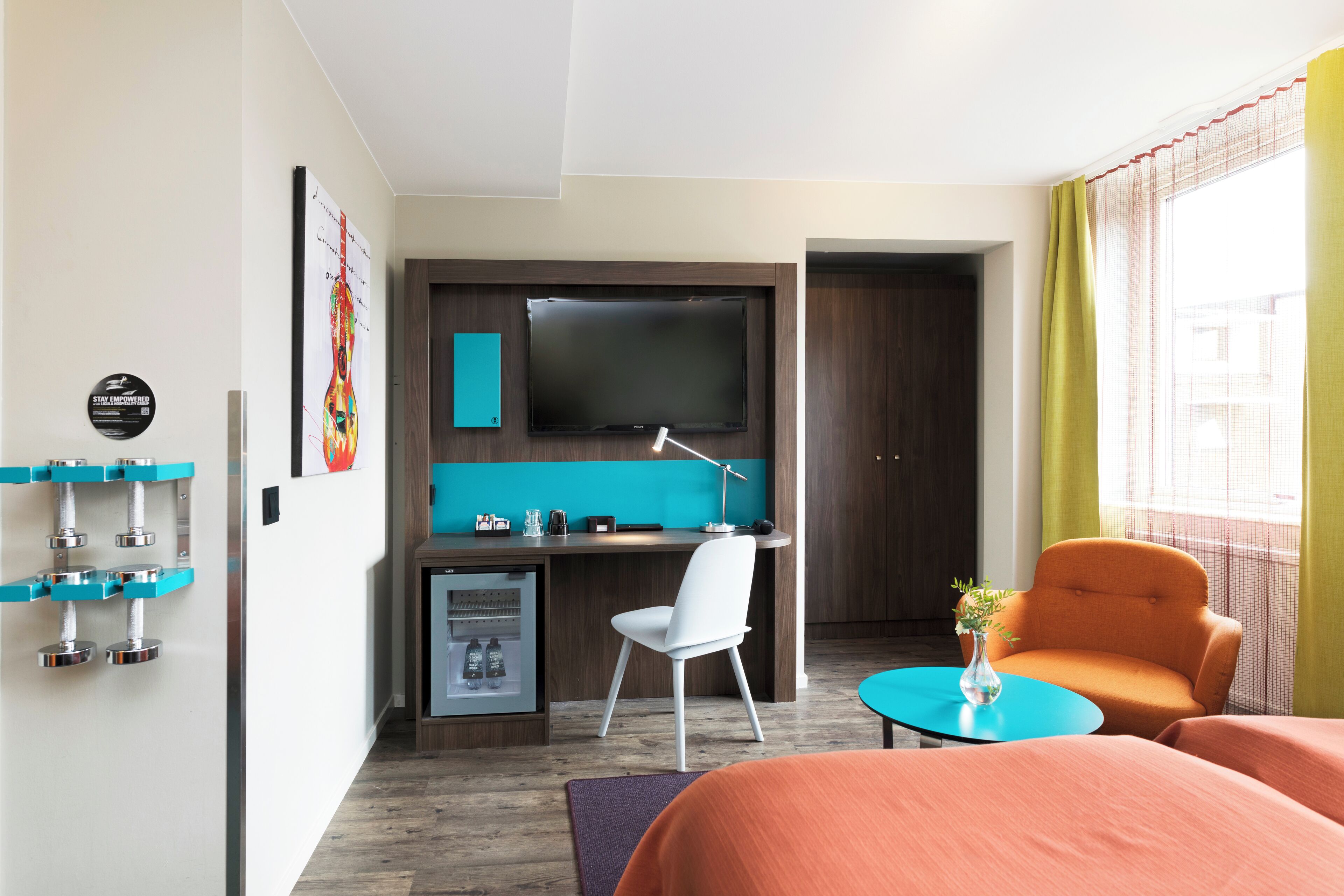A hotel room with a bed, a desk with a TV, a small fridge, a chair, and a window with curtains.