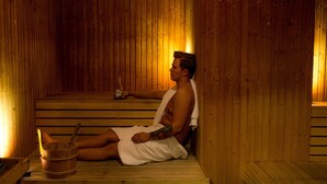 A wooden sauna with a person sitting on a bench, a bucket, and a bottle. - ProfilHotels Savoy (Jonkoping)