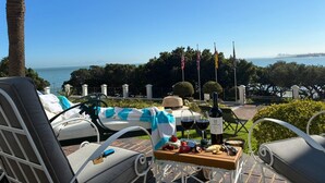 A terrace with outdoor seating, a table with wine glasses and snacks, and a view of the sea and city skyline.