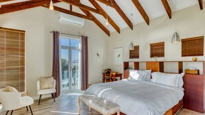 A spacious bedroom with a large bed, wooden beams, a bench, a chair, and a view through a glass door.