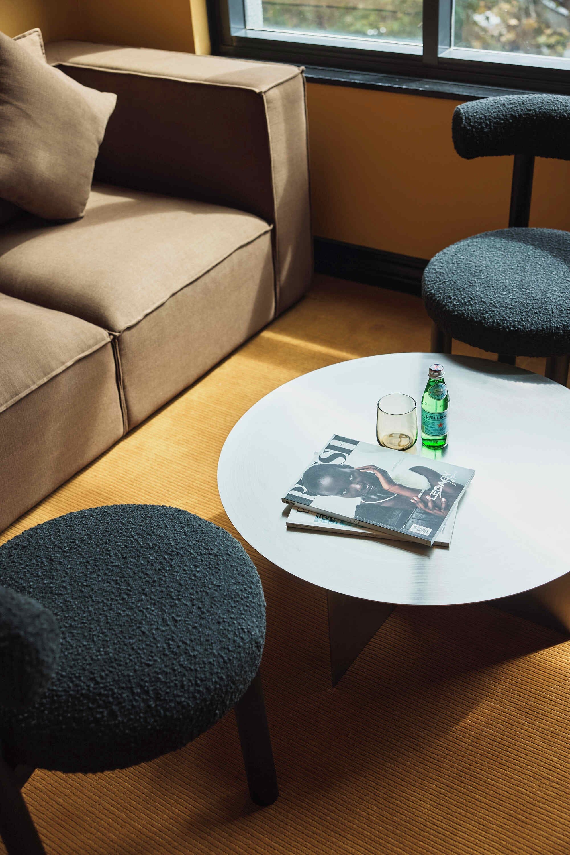 A modern living room with a beige sofa, two dark chairs, a round white table with a magazine, a glass, and a bottle.