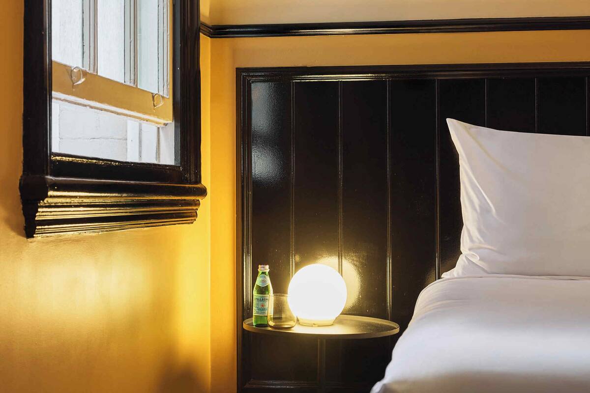 Hotelkamer met een bed, nachtkastje met lamp, fles water en raam met jaloezieën.