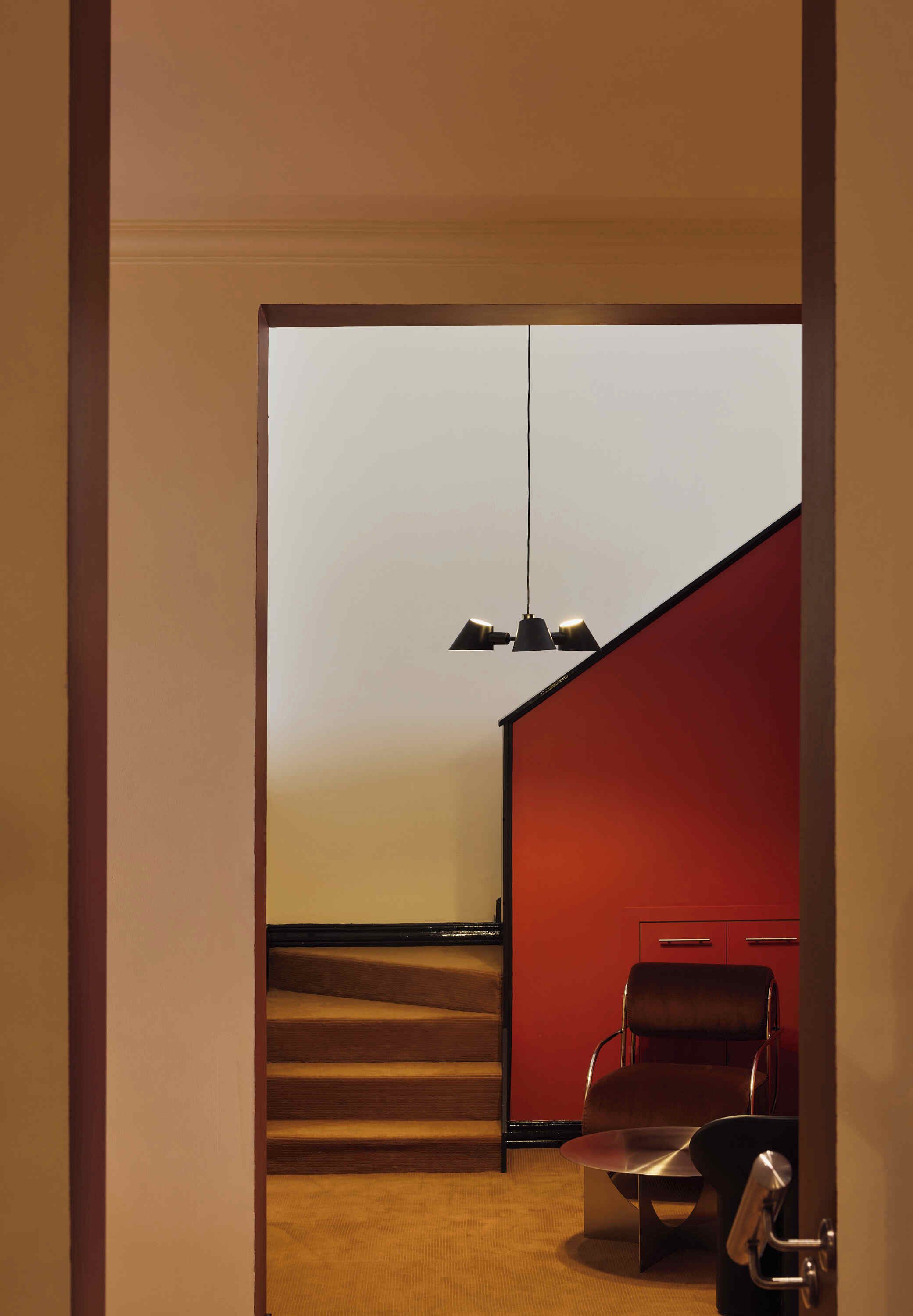 Um interior moderno com uma escada que leva a uma parede vermelha, uma cadeira preta e um lustre pendurado.