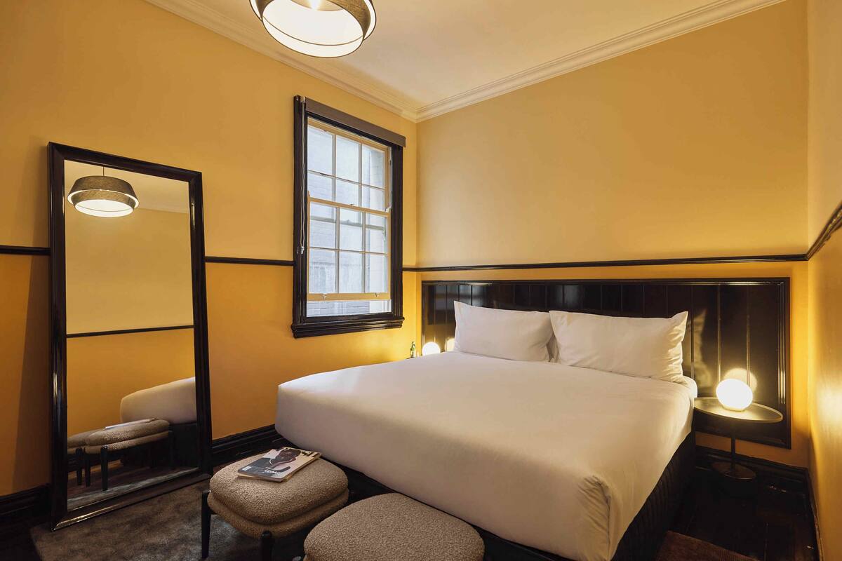 A hotel room with a large bed, a bedside table with a lamp, a window, a mirror, and a small stool.