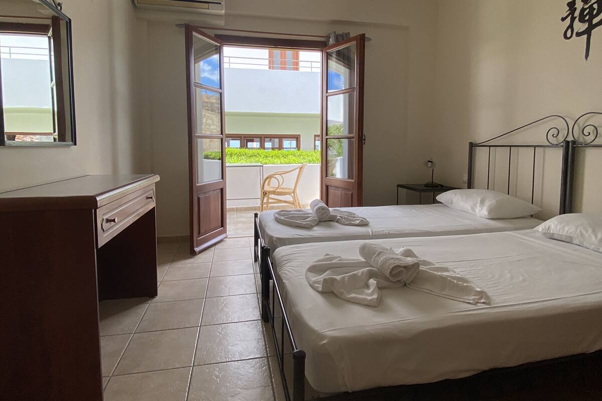 A hotel room with two beds, a desk, and a balcony with a view.