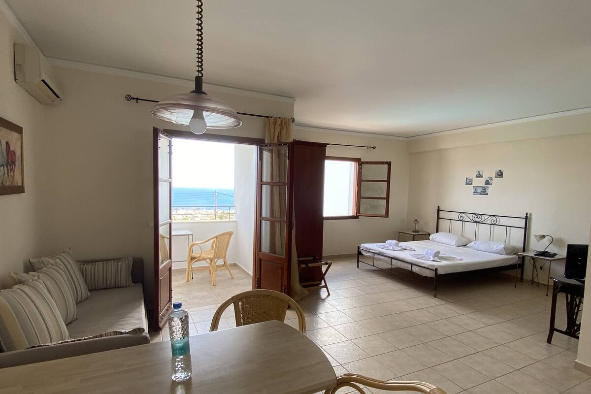 A hotel room with a bed, a sofa, a dining table with chairs, a TV, and a view of the sea.