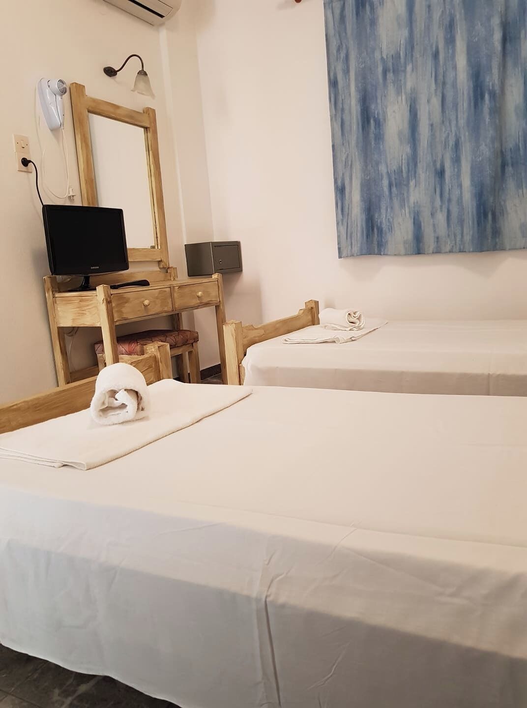 A hotel room with two beds, a wooden desk with a computer, a hairdryer, and a wall-mounted lamp.