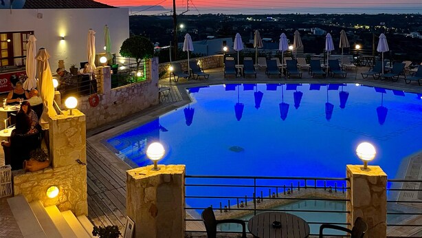 A swimming pool with lounge chairs and umbrellas, surrounded by stone walls and a view of the city at sunset. - Amazones Village Suites (Hersonissos)