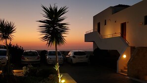 A modern building with a staircase leading to the upper floor, surrounded by parked cars and a palm tree. - Amazones Village Suites (Hersonissos)