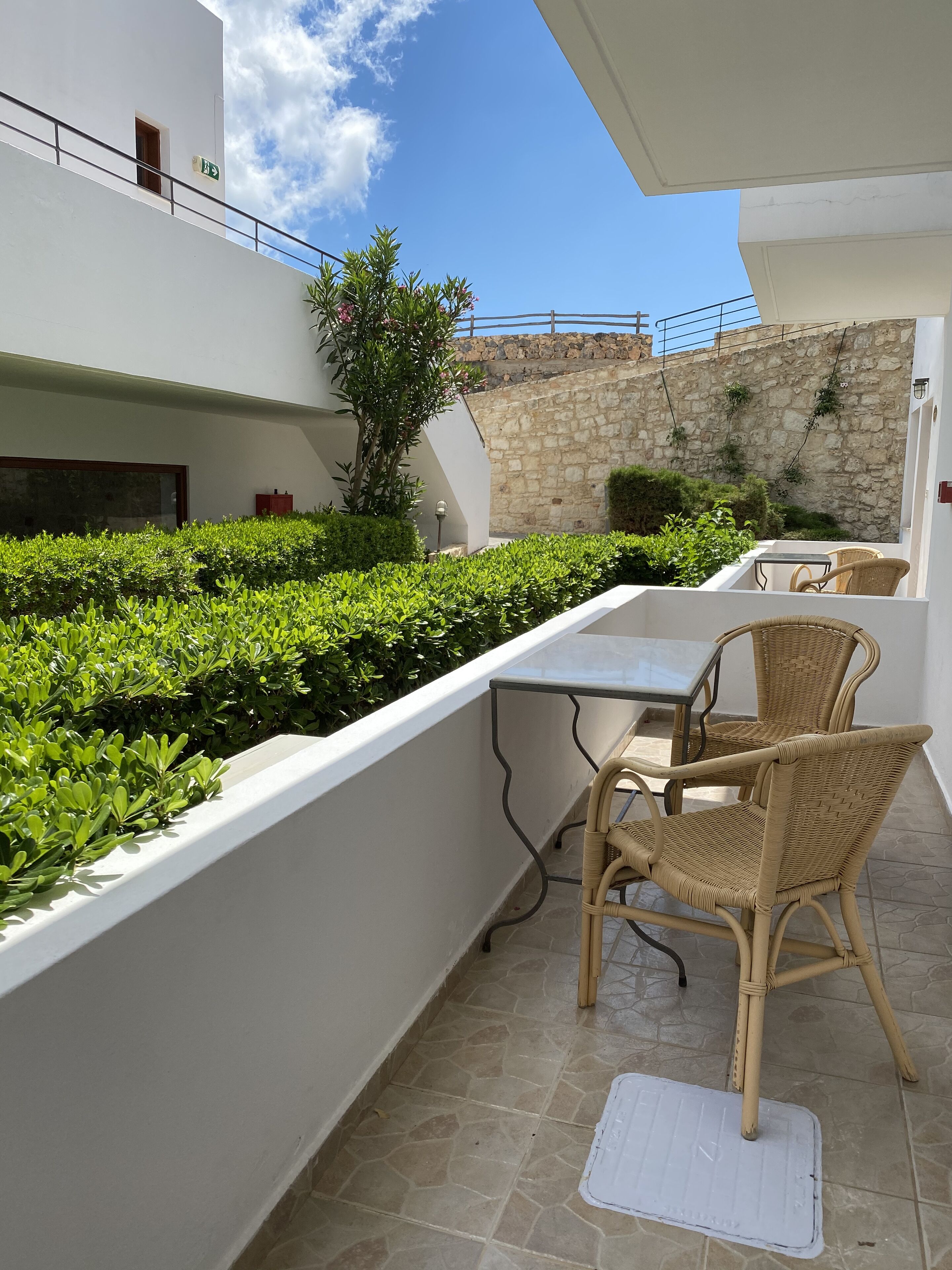 Un balcon agrémenté d’une table et de deux fauteuils en osier, entouré d’arbustes soigneusement taillés et d’un mur en pierre.