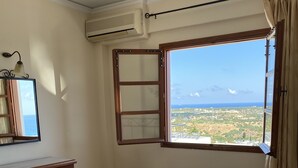 A hotel room with a bed, a desk, a mirror, and a window with a view of the landscape. - Amazones Village Suites (Hersonissos)