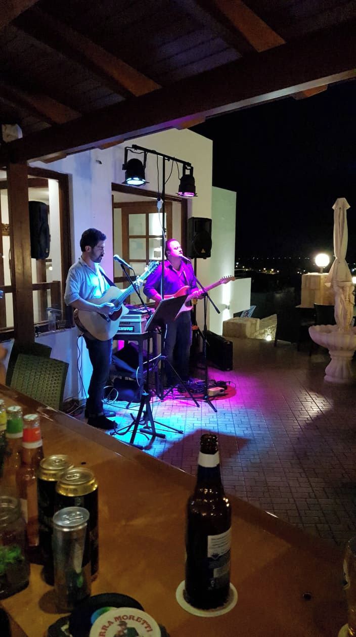 An outdoor patio with a musician playing a guitar and a keyboard, a table with drinks, and a statue.