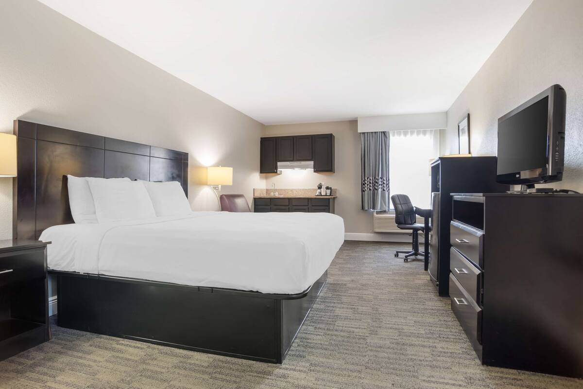 A hotel room with a large bed, a desk with a chair, a television, and a kitchenette area.