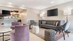 Un salon moderne avec un canapé gris, des fauteuils violets, une table basse et un pouf à motifs.