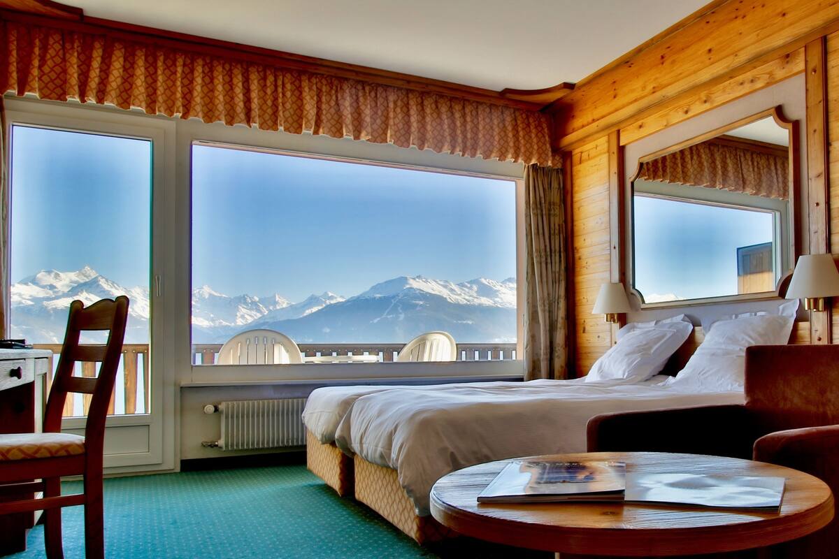 Ein Hotelzimmer mit Bett, Sessel, Tisch und großem Fenster mit Blick auf die Berge.