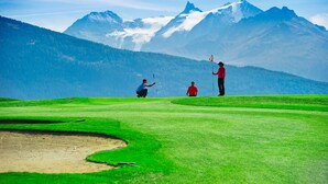 Een golfbaan met een berg op de achtergrond, een vlaggenstok en een golfer in een rood shirt.
