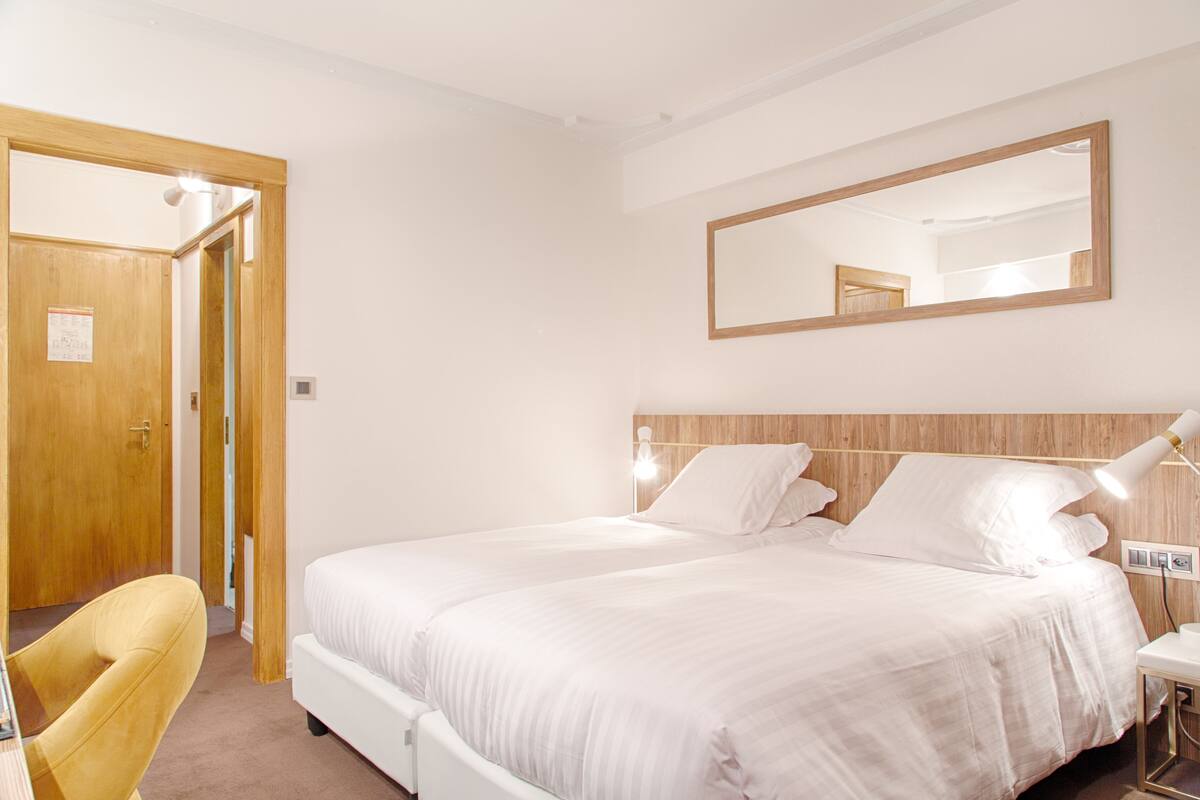 Ein Hotelzimmer mit Doppelbett, Holz-Kopfstück, Nachttischlampe, einem Sessel und einer Tür zu einem anderen Raum.