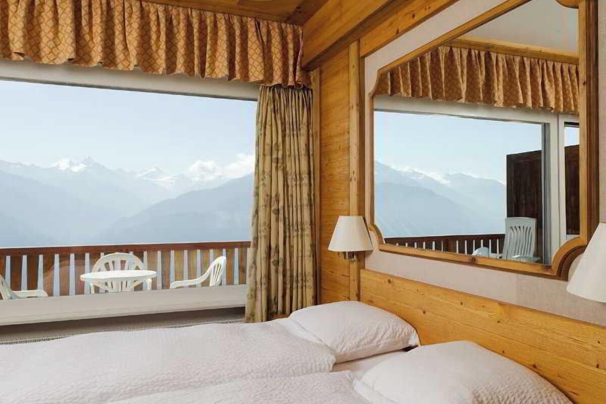 Habitación de hotel con cabecera de madera, ventana grande con vistas a montañas, cama con ropa blanca y una pequeña mesa con sillas.