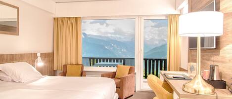 A hotel room with a bed, a desk, a chair, a lamp, and a view of mountains through the window.