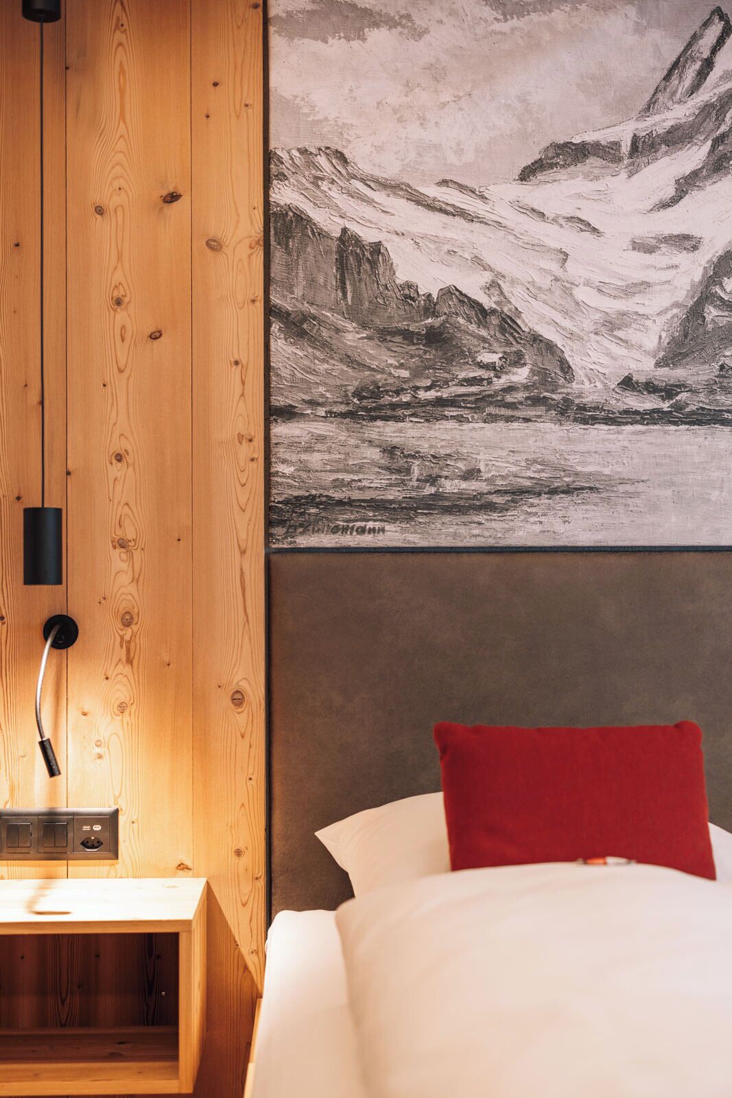 A bedroom with a wooden headboard, a bedside table with a lamp, a red pillow, and a large black-and-white mountain landscape painting on the wall.