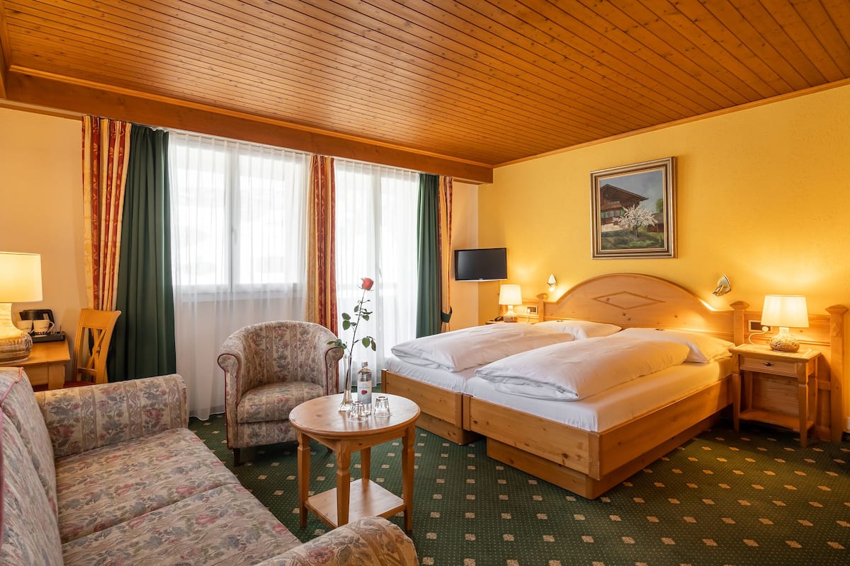 A hotel room with a wooden ceiling, two beds, a sofa, a chair, a small table with a vase, and a painting on the wall.