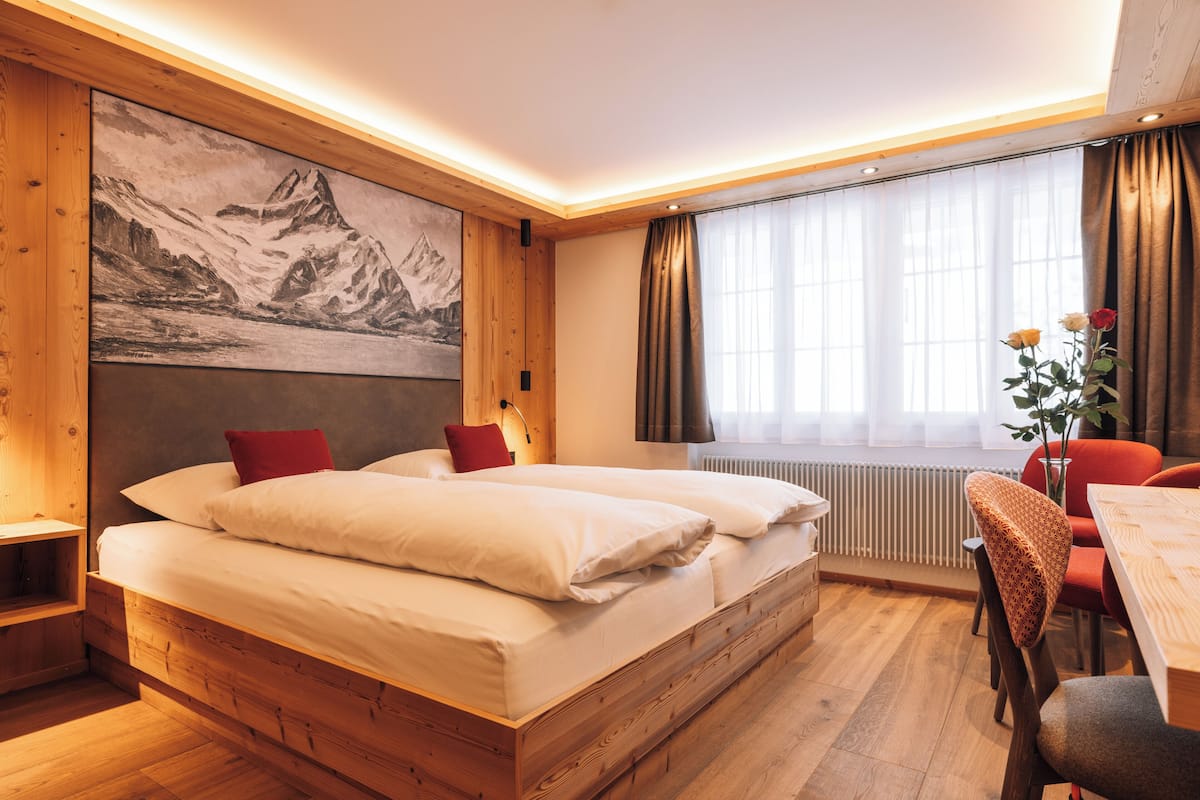 A bedroom with a wooden bed, a large painting of mountains, a window with curtains, and a table with chairs.