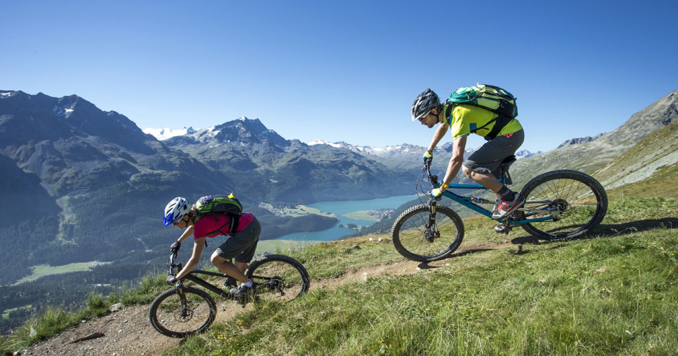 Dos ciclistas de montaña en un sendero con un lago y montañas al fondo.