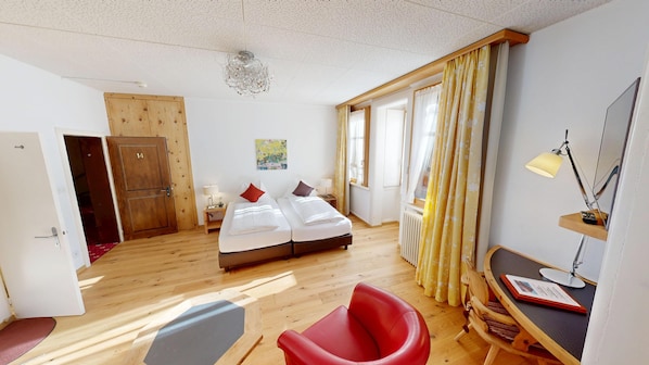 Una habitación de hotel con suelo de madera, una cama con ropa de cama blanca, una silla roja, un escritorio con una lámpara, una ventana con cortinas amarillas y una puerta de madera con el número 14.