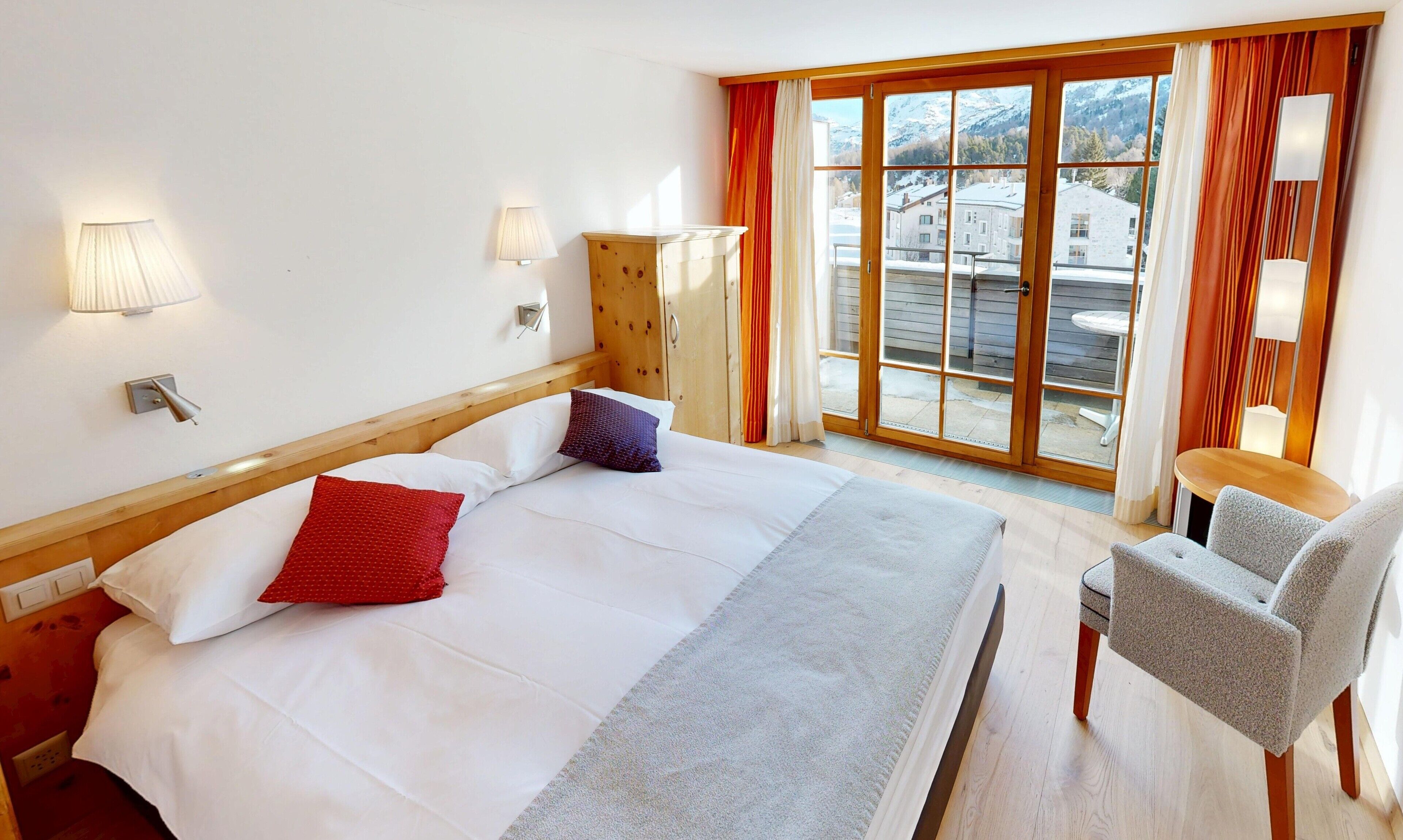 Une chambre d’hôtel avec un grand lit, deux lampes de chevet, une armoire en bois, une chaise et une vue sur les b âtiments et les montagnes par la fenêtre.