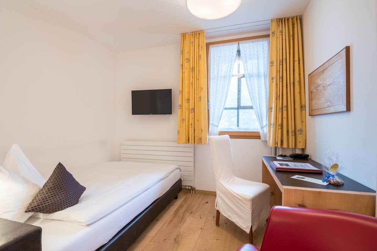 Ein Hotelzimmer mit Bett, Schreibtisch, Stuhl, Fernseher und einem Fenster mit Vorhängen.