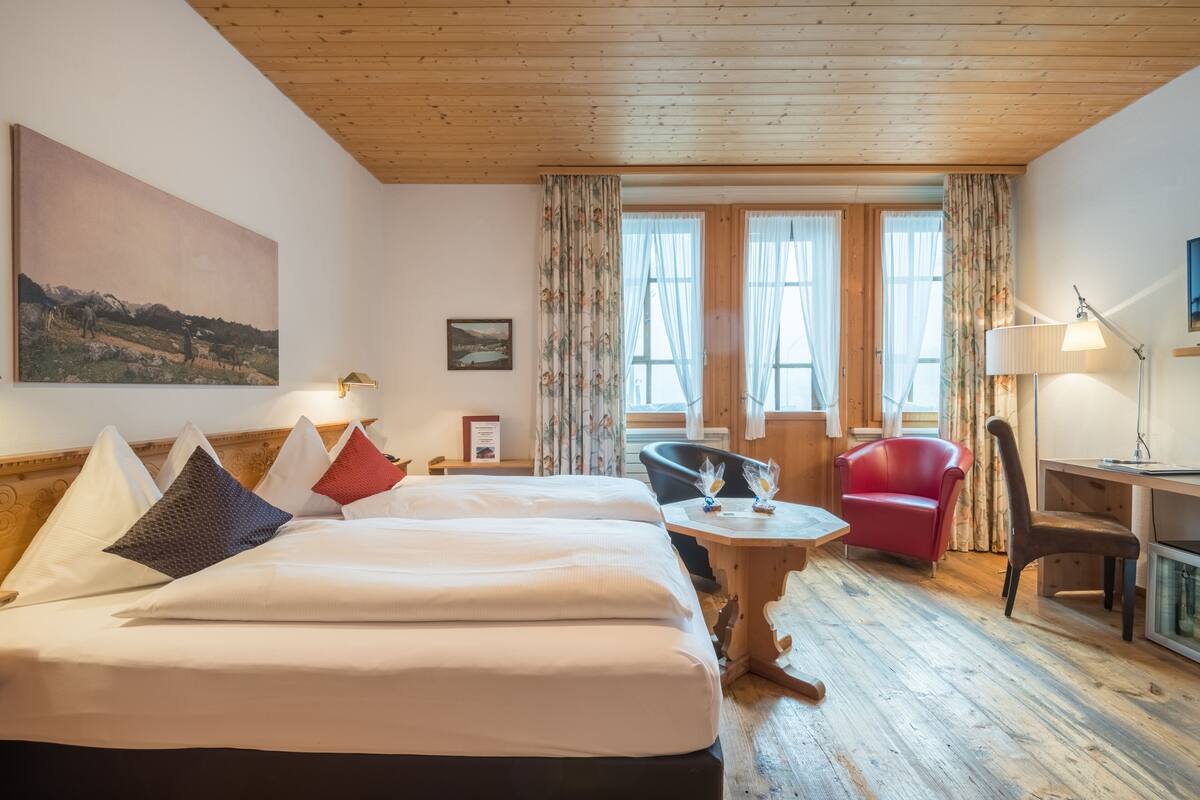 Ein Hotelzimmer mit Holzboden, einem großen Bett mit vielen Kissen, einem Schreibtisch mit Lampe, einem roten Sessel und Blick auf eine Landschaft durch das Fenster.