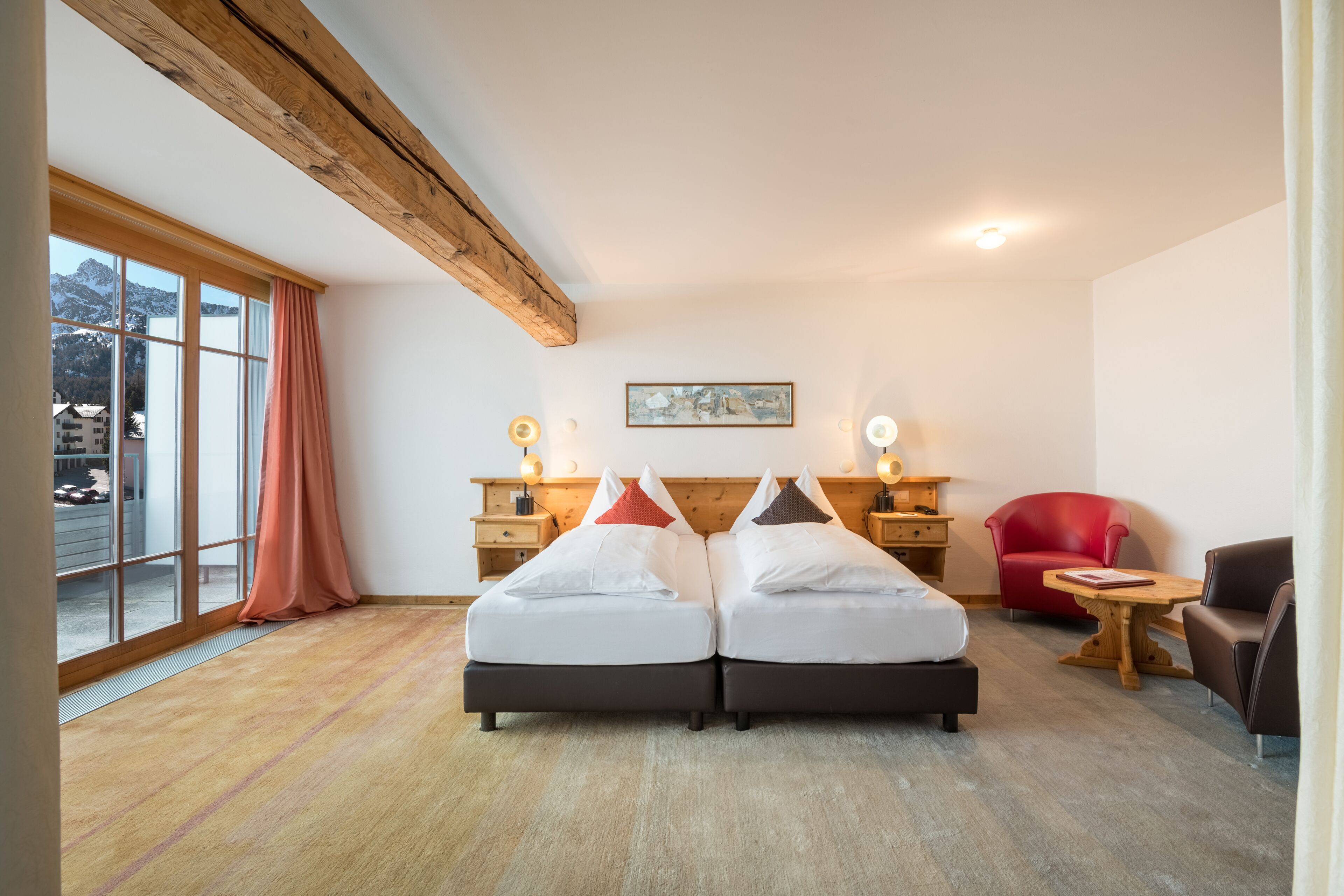 Ein Zimmer mit zwei Betten, Holzbalken, einem großen Fenster mit Vorhängen, einem roten Stuhl und einem Tisch.