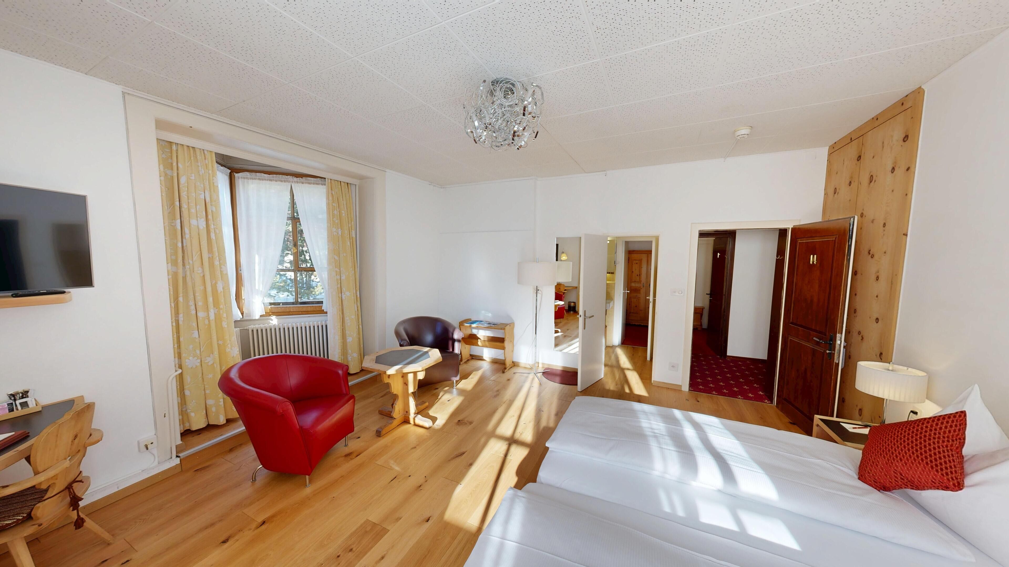 A hotel room with a bed, a red armchair, a small table, a lamp, a TV, and a window with curtains.