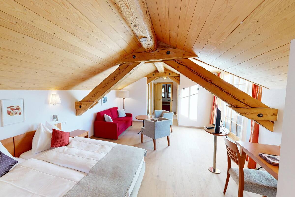 Ein geräumiges Dachzimmer mit hoher Decke, Holzbalken, einem roten Sofa, einem weißen Sofa, einem blauen Sessel und einem Esstisch.