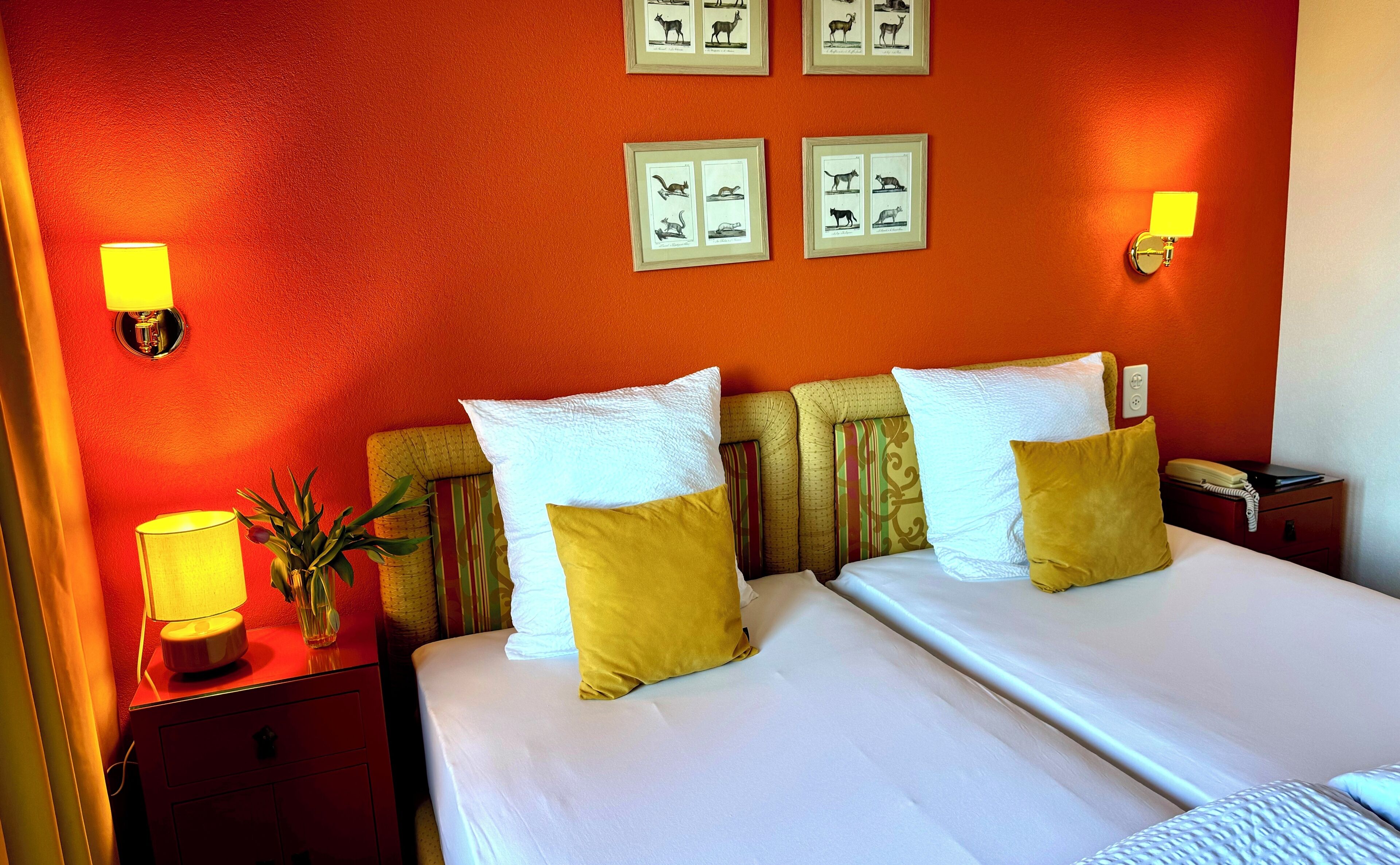 Quarto de hotel com duas camas, mesa de cabeceira com abajur, telefone e ilustrações de animais emolduradas na parede.