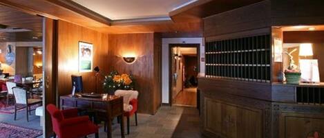 A hotel lobby with a wooden reception desk, a seating area with chairs and a rug, and a wall-mounted light fixture.