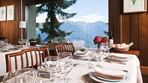 A dining table set with plates, glasses, and cutlery, with a mountain view through a window. - Hôtel Résidence de la Forêt (Crans-Montana)