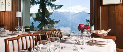 A dining table set with plates, glasses, and cutlery, with a mountain view through a window.
