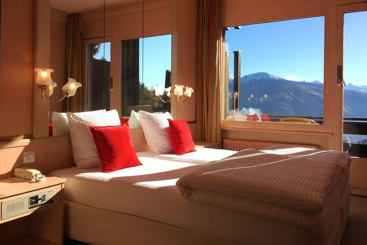 A hotel room with a large bed, a bedside table with a telephone, and a window offering a mountain view.