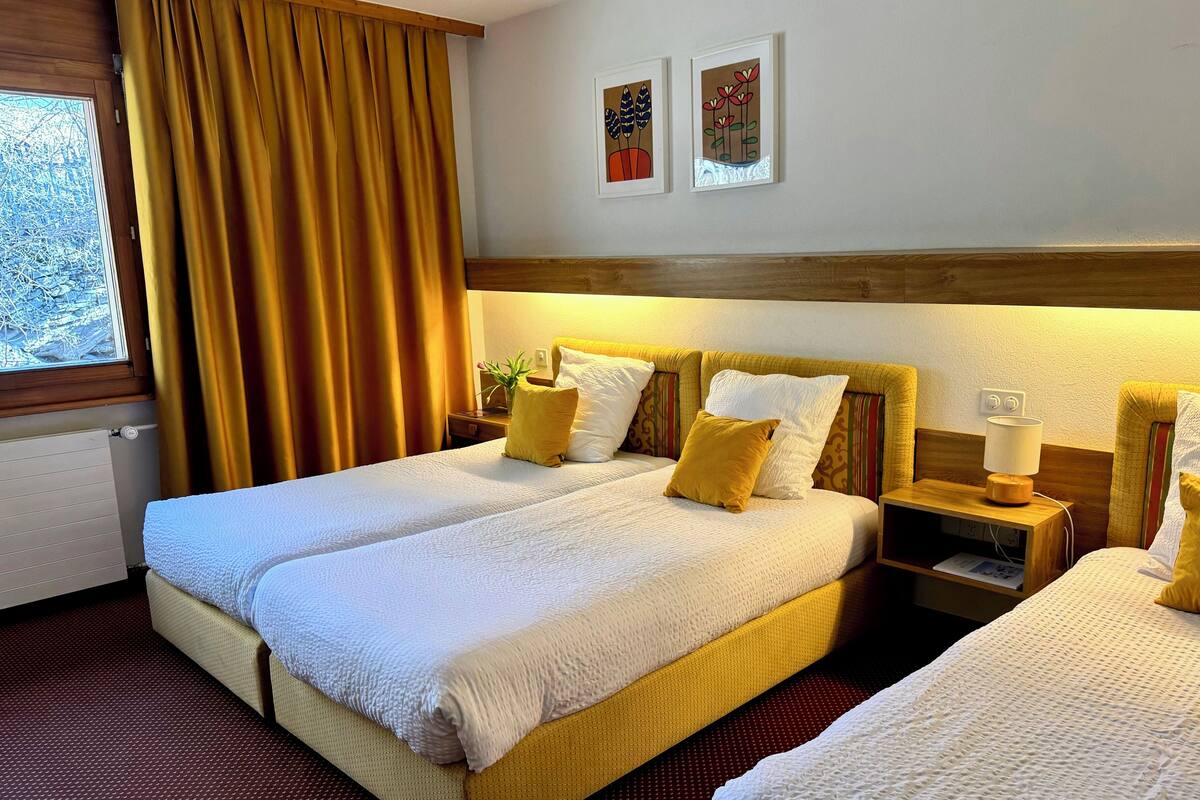 A hotel room with two beds, a window with curtains, a nightstand with a lamp, and two wall-mounted paintings.