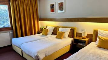 A hotel room with two beds, a window with curtains, a nightstand with a lamp, and two wall-mounted paintings.