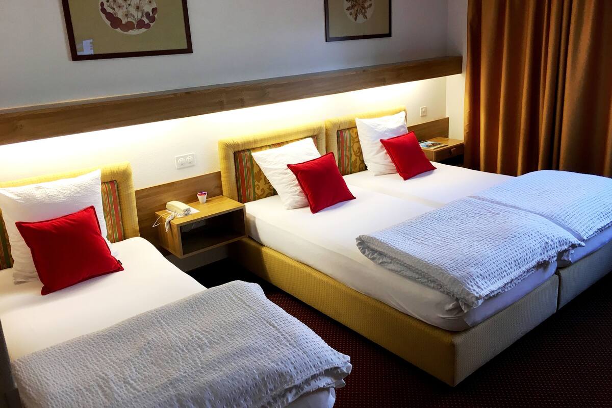 A hotel room with two beds, each with white linens and red pillows, a wooden headboard, bedside tables with lamps, and framed artwork on the walls.