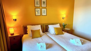 Quarto de hotel com duas camas, mesa de cabeceira com abajur, iluminação embutida na parede e ilustrações botânicas emolduradas.