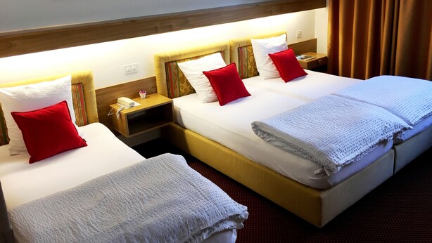 A hotel room with two beds, each with white linens and red pillows, a wooden headboard, a nightstand with a phone and a small table, and framed artwork on the wall. - Hôtel Résidence de la Forêt (Crans-Montana)