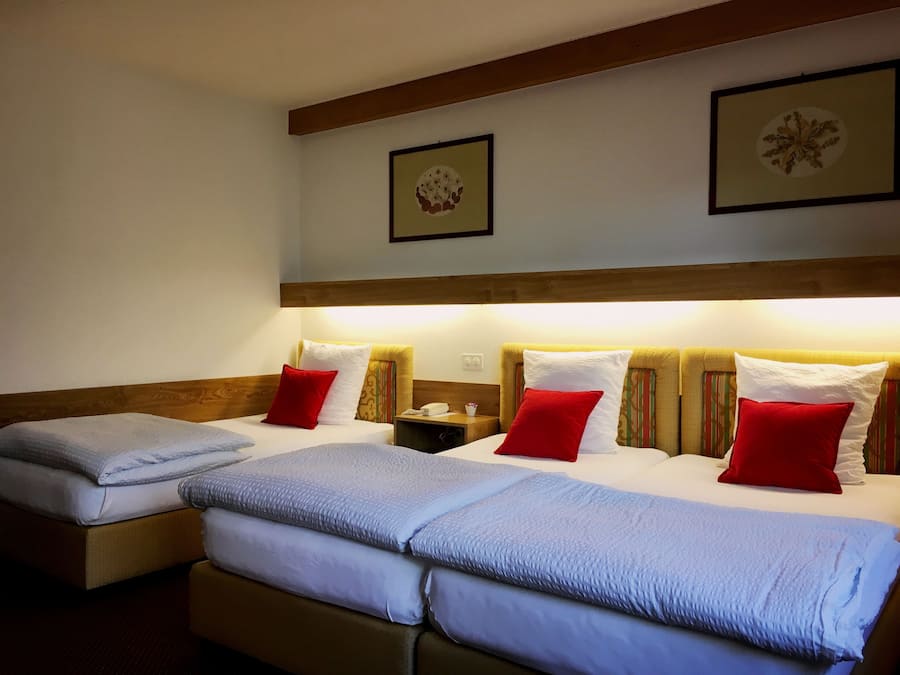 A hotel room with two beds, each with a headboard, white linens, and red pillows. There are framed pictures on the wall above the beds.