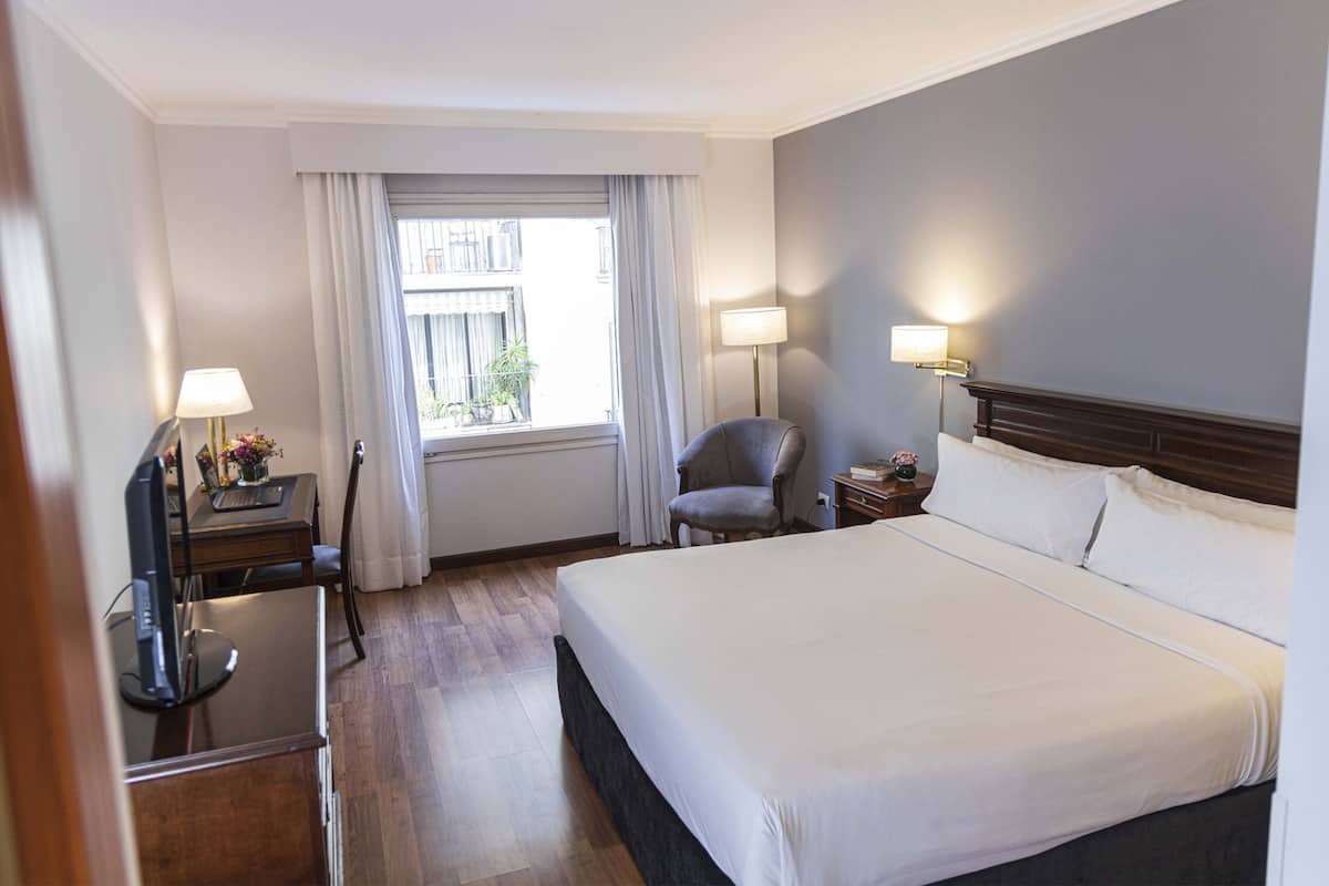 A hotel room with a large bed, two bedside lamps, a chair, a desk with a computer, and a window with curtains.
