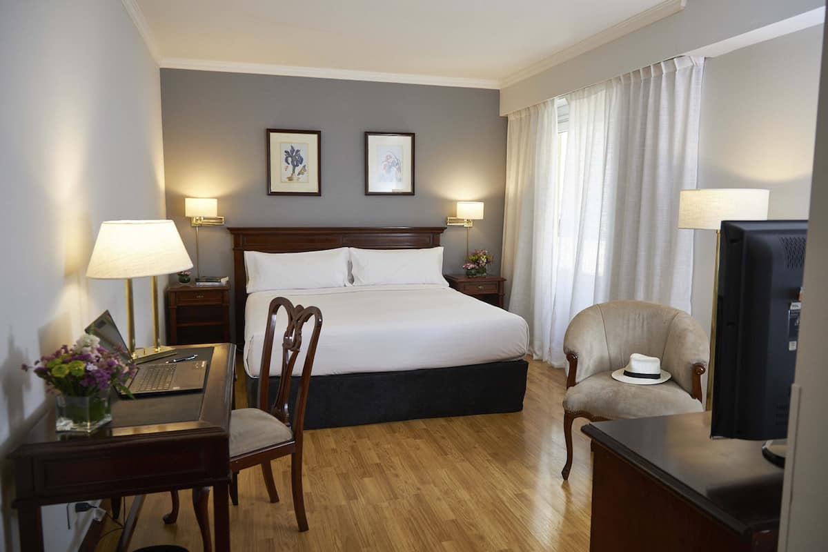 A hotel room with a bed, a desk with a laptop, a chair, a television, and two framed pictures on the wall.