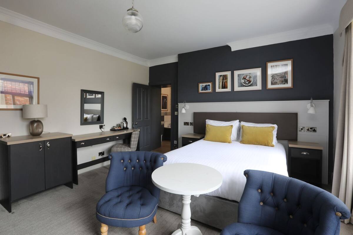 A modern hotel room with a large bed, two armchairs, a small white table, and a desk with a chair. There are framed pictures on the wall and a door leading to another room.