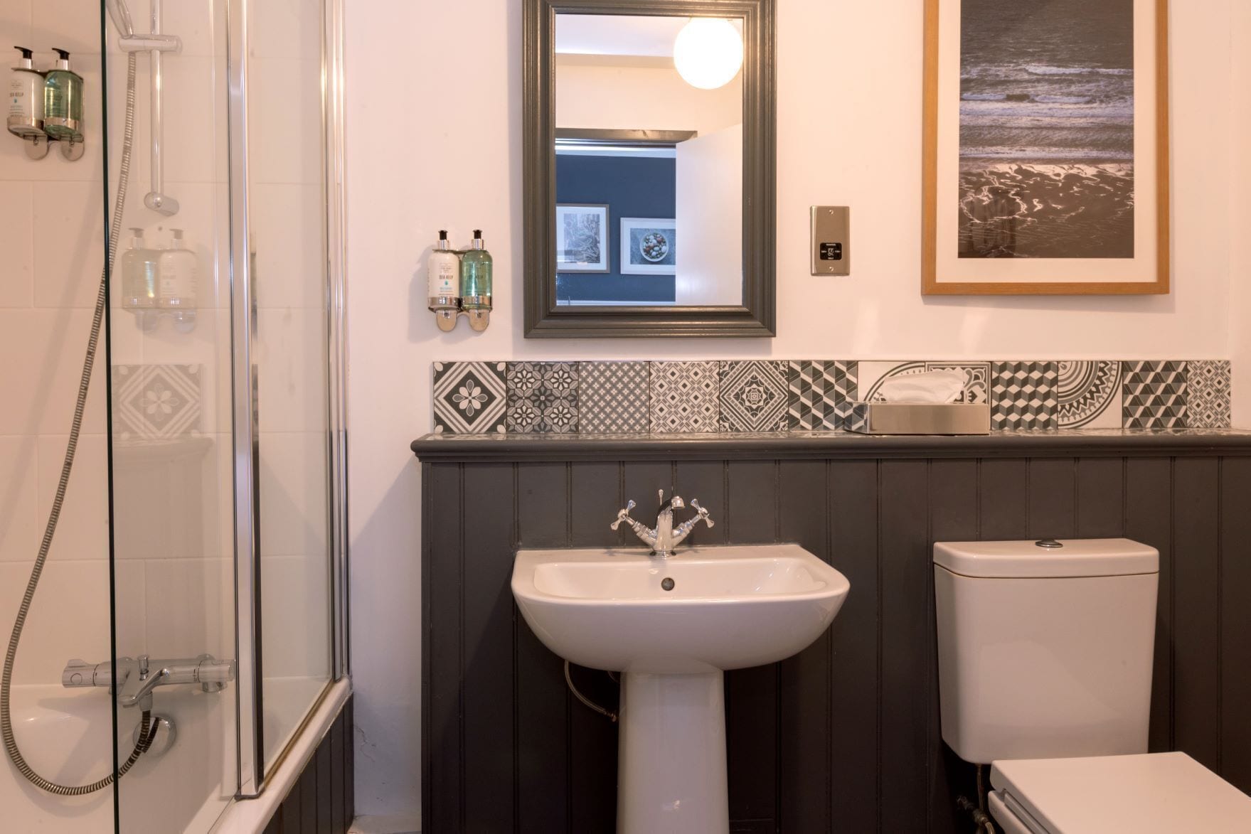 Un baño con ducha, lavamanos e inodoro. Hay azulejos decorativos en la pared y un cuadro enmarcado sobre el lavamanos.