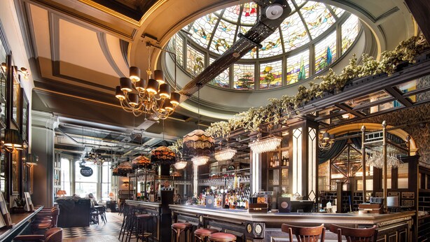 Un bar con techo alto, candelabros ornamentales y un amplio ventanal de vitral.